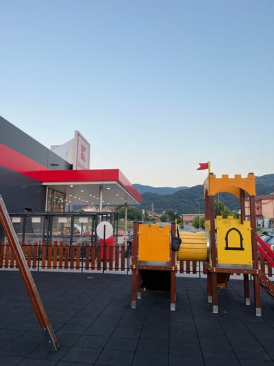 This image captures an outdoor view in Petrich, Bulgaria, featuring a playground in the foreground and a Kaufland supermarket in the background. The playground equipment includes a castle-themed play structure with yellow panels, a red flag on top, and a tunnel slide. A swing set frame is also visible on the left. The ground is covered with black rubber safety tiles. Behind the playground, a wooden fence separates it from the supermarket's entrance area, where shopping carts are lined up. A portion of the Kaufland store's exterior, with its distinctive red and grey facade and illuminated signage, is prominent. The scene is set against a backdrop of rolling, tree-covered hills under a clear, pale blue sky, suggesting it is late afternoon or early evening.