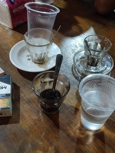 This is a close-up, eye-level shot of a wooden table with several glasses and a cigarette pack. The scene appears to be set in an informal establishment, possibly a cafe or warung, in Keudematangkuli, Indonesia.
In the foreground, a glass contains dark liquid, likely coffee, with a spoon resting inside. To the right, another glass is filled with clear water. Behind these, a small white saucer holds a clear glass with a dark residue at the bottom, possibly from a previous drink. Stacked on top of this glass is a clear plastic cup, empty. To the left of the coffee glass, a pack of cigarettes is visible, with partial text including "MO" and "10" along with Indonesian text that seems to be a warning about health.
The lighting suggests it could be daytime, with shadows cast on the table, indicating diffused light. The wooden table has a worn texture, adding to the casual atmosphere. The overall mood is relaxed and everyday, implying a moment of pause or a casual meeting. The presence of coffee and cigarettes suggests a common break or social activity. No people are visible in the frame.