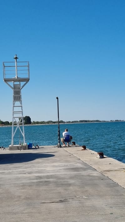A person is fishing on a pier in Limani Litochorou, Greece. The person is sitting in a blue chair facing the water. A white tower is to the left of the person. Fishing rods and a blue bucket are on the pier. The water is blue, and the sky is clear. The pier is made of concrete. Trees and buildings are visible in the distance on the shore.