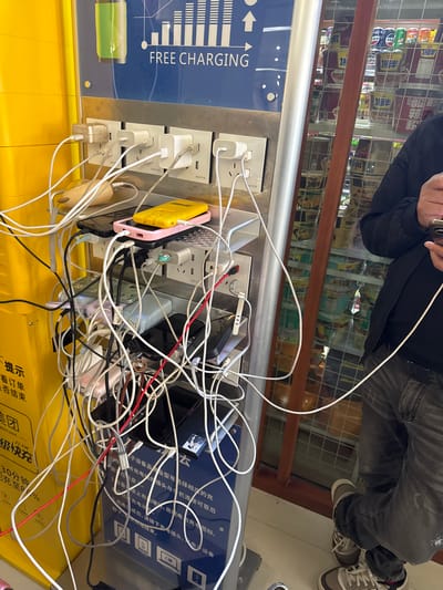 This image captures a busy public charging station in Dunhuang City, China, likely within a retail or public space. A person, whose upper body and legs are visible, stands near the station, possibly engaging with a mobile device. They are dressed in a black jacket, dark jeans, and sneakers.

The main subject is a "FREE CHARGING" kiosk, which is in heavy use. Numerous white, black, and red charging cables are tangled together, connecting various electronic devices—including a yellow power bank or phone and a pink-cased phone—to multiple power outlets. Several white power adapters are plugged into the vertical outlets on the station.

The charging station features a blue panel with prominent English text "FREE CHARGING." Chinese text on the panel includes "提示" (Tip), "美团" (Meituan), "22.5W 级快充" (22.5W Super Fast Charge), and "30分钟 充至80%" (30 minutes charge to 80%), indicating rapid charging capabilities. Further instructions in Chinese describe the charging process, advising users to connect securely and unplug after use. Small text on the outlets reads "复路科技" (Fulu Technology).

In the background, behind a wooden railing, a refrigerated display case is visible, stocked with a variety of canned beverages, including brands like Pepsi, "康师傅" (Master Kong), and "旺仔" (Want Want), suggesting the setting is a convenience store or a similar establishment. The scene conveys a sense of practicality and the common modern need for on-the-go device charging.