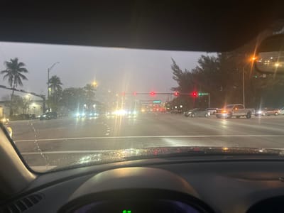 The image depicts an urban intersection in Doral, Florida, viewed from the interior of a stationary vehicle during conditions of precipitation or a recently wet environment. Water droplets are visible on the vehicle's windshield and dashboard.
The viewer's vehicle is stopped at a red traffic signal. Multiple red lights are illuminated for the viewer's direction of travel, while green lights are displayed for cross-traffic. A green street sign, affixed to a traffic light pole on the right side of the intersection, clearly indicates "NW 36 ST," identifying the intersecting road.
The asphalt roadway is wet and reflective, with visible white lane markings. Numerous vehicles are present at the intersection. A light-colored pickup truck is prominently visible in a lane on the cross street, its headlights illuminated. Other sedans and vehicles are visible with active headlights, either stopped or proceeding through the intersection. The environment includes multiple palm trees and dense foliage lining the roadsides. Overhead, the sky is grey and overcast, suggesting dusk, dawn, or heavy cloud cover. Streetlights and vehicle lights are illuminated. No individuals are discernible.