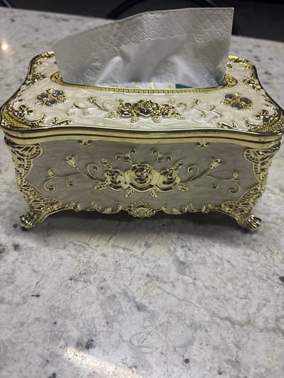 A decorative, rectangular tissue box with an ornate gold trim sits on a speckled white countertop. The tissue box is predominantly cream-colored with a swirling pattern and features raised gold embellishments, including floral designs and what appear to be small, clear stones. The top of the box has a gold-plated opening from which white tissues are protruding. The box rests on four curved gold legs. The background shows a blurred dark surface and the countertop exhibits natural stone patterns with varying shades of gray, white, and dark flecks. There are no people visible in the image. The image appears to have been taken indoors. The location is Ghukasavan, Armenia.