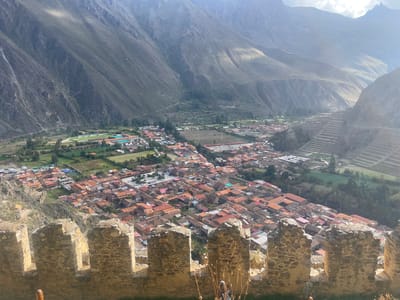 The scene is a high-angle, daytime view of the city of Ollantaytambo, Peru, framed by an ancient stone wall in the foreground. The wall, constructed of irregular, light-colored stones, runs horizontally across the lower portion of the image. Behind the wall, a dense arrangement of buildings with predominantly red-tiled roofs spreads out into the middle ground. Beyond the buildings, a green field area is visible with a sports complex. In the background, towering mountains dominate the landscape, their slopes covered in varying degrees of vegetation. The sky is partially obscured by the mountain peaks and appears hazy due to atmospheric conditions. The overall impression is one of a historic settlement nestled within a rugged mountainous region.