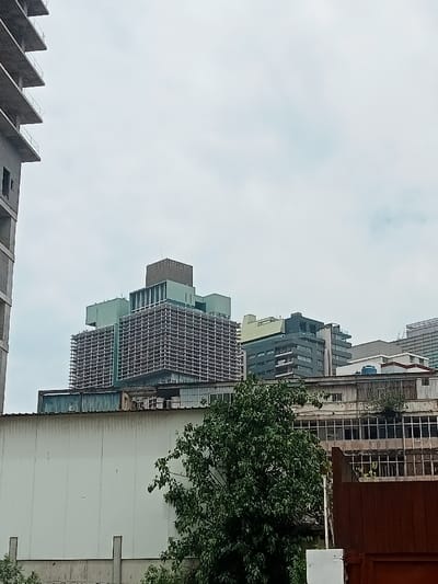 This image captures an urban landscape in Luanda, Angola, on an overcast day. The scene is dominated by a mix of modern and older buildings under a uniformly grey, cloudy sky.
In the left foreground, a tall, partially constructed concrete building with exposed floor slabs and rebar rises vertically, indicating ongoing development. To its right, in the mid-ground, stands a distinctive, multi-story building featuring a prominent gridded facade, possibly a Brutalist or mid-century modern design. The upper sections of this building incorporate lighter green or teal-colored panels and a darker, box-like structure on top.
Further to the right and in the background, several other modern buildings are visible, some with dark grey or black facades, balconies, and accents of the same green/teal color seen on the central building. Below these, a long, low-rise structure stretches across the mid-ground, characterized by a corrugated metal roof and numerous windows or openings along its facade. The left portion of this lower structure presents a plain, light-colored wall.
A lush, leafy green tree occupies the lower mid-ground, partially obscuring the base of the lower buildings. In the far lower right, a dark brown, possibly metallic, fence or gate is visible.
There are no people or specific activities taking place, and no discernible text is visible. The time of day appears to be daytime, though the heavy cloud cover prevents any direct sunlight, contributing to a muted and somewhat industrial atmosphere.