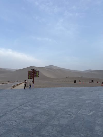 The image depicts a view of Mingsha Mountain in Dunhuang City, China. A large sand dune rises adjacent to a paved walkway. Along the dune's base is a series of wooden posts, potentially forming a barrier or guide. A signpost with multiple directions is prominent in the foreground. Several people are visible near the signpost and along the dune, suggesting tourist activity. The walkway extends into the distance. The sky is overcast with light clouds.