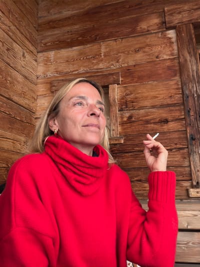 A solo adult female is depicted in a medium shot, seated in a wooden structure in La Clusaz, France. She has blonde hair and wears a bright red cowl-neck sweater with ribbed cuffs and gold-toned hoop earrings. Her head is tilted slightly upwards, and her gaze is directed towards the upper left, exhibiting a neutral expression. Her right hand is raised, holding a lit white cigarette with a visible filter between her index and middle fingers. The background features aged, horizontally-oriented wooden plank walls, characterized by natural grain and knots. To the right of the subject's head, a small, dark rectangular opening, framed by vertical wooden beams, is integrated into the wooden paneling.