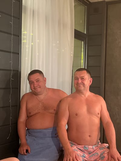 Two shirtless men are posing for a photo in what appears to be an outdoor setting. The man on top is wearing pink patterned shorts, while the man below is wearing blue shorts. The location includes a window and a white curtain. The background also has some string lights. This appears to be a candid, informal photograph. There is no specific information linking the image to the city of Новая Чернушка, Russia.