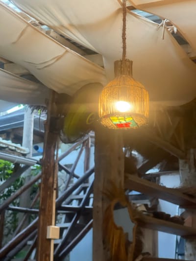 This image, captured in Michamvi, Tanzania, depicts an illuminated indoor or semi-outdoor space characterized by rustic construction. A central hanging light fixture, crafted from woven natural fibers in a bottle-like form, emits bright, warm light from an exposed bulb. The lower portion of the fixture features square panels of colored glass or plastic in red, yellow, and green. The light is suspended by a wrapped cord from an overhead structure covered by draped, off-white fabric panels. To the left and slightly behind the light, a lime-green wall-mounted oscillating fan is partially visible. The environment features multiple unprocessed wooden posts and crossbeams, forming a multi-tiered structural framework. A wooden staircase is discernible ascending in the background, with a light blue wall section beyond it. A small, white electrical box is affixed to a wooden post in the lower-left quadrant, and a decorative, irregularly shaped wooden mirror or cutout adorns a vertical wooden beam on the right. No individuals are present in the scene.