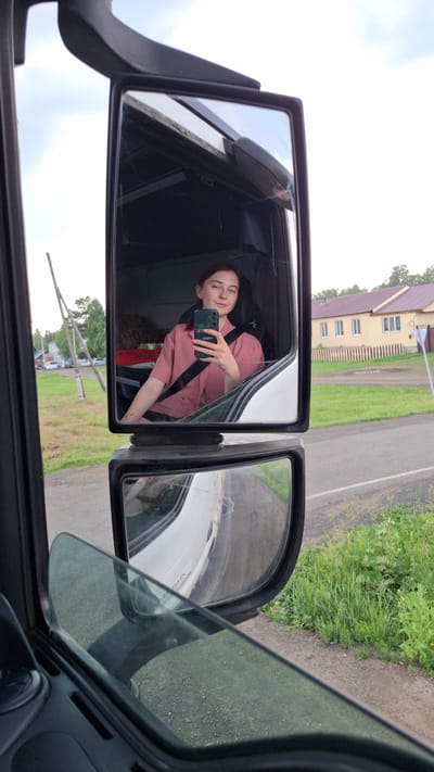 A young woman with short red hair is sitting in the driver's seat of a truck, taking a selfie with her phone. She is wearing a pink collared shirt and has a seatbelt on. The truck's large side mirror reflects her and the interior of the cab. Outside the truck, visible in the mirror, is a rural scene with a road, grassy verge, trees, and a two-story building with a yellow facade and a red roof. The lower mirror reflects the road and the side of the truck as it moves. The scene appears to be in a rural area outside of Nizhny Ingash, Russia.
