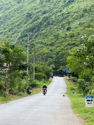 The image depicts a rural road in Cán Tỷ Commune, Vietnam, flanked by lush green, tree-covered mountains. The road is paved and curves gently into the distance. On the left side of the road, there are power lines supported by utility poles, and some residential structures are visible amidst trees. On the right side, more vegetation lines the road, and a white milestone marker indicates "Km 62 TRANG KIM Km 62." A person on a scooter is riding towards the viewer on the right side of the road. Another scooter is parked on the left shoulder. Further down the road, a group of people and several vehicles, including what appears to be a small truck, are gathered. A bird is perched on the edge of the road near the milestone marker. The overall scene suggests a tranquil, natural environment with signs of local life and infrastructure.