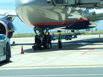 The image shows an airplane on the tarmac at Cam Ranh International Airport in Cam Ranh, Vietnam. The nose of the aircraft is prominent, with a blue and red engine cowling visible on the left. The underside of the plane's fuselage, which is silver with a red stripe, is also visible. Ground equipment, including a blue baggage tractor, is parked near the aircraft. Two individuals, likely airport personnel, are standing near the tractor. One is wearing a blue uniform and the other a high-visibility green vest. A traffic cone is positioned on the tarmac. The sky is a light blue, and the ground is paved with concrete. The overall scene depicts a typical airport environment.