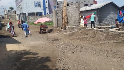 The image shows a dirt road in Rubavu, Rwanda, at coordinates -1.664 latitude and 29.276 longitude. There are five people walking on the road: a woman in a red and blue dress, two men in blue shirts, a woman in a yellow dress, and a man in a turquoise shirt. One of the men on the road is pushing a handcart. There is a pink umbrella on the handcart, a motorcycle parked on the road, and a gray building to the right. The building is made of corrugated metal and has a concrete foundation. There are also some wooden beams and a pile of dirt in front of the building.  The road is lined with green plants and a few trees, while the sky is a clear blue. The street is empty except for the individuals walking and the motorcycle.