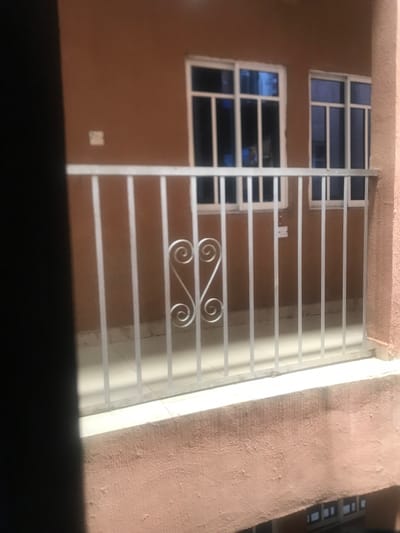 The image shows a close-up view of a window with a white metal grate in front of it. The grate is made of horizontal bars and features a decorative design at the bottom. The window is divided into two sections, with the top section having a small space between each pane of glass and the bottom section having a larger space between each pane. The window is on a brown wall, with a light brown trim. The building is located in Umudike, Nigeria, at the coordinates 5.471° N, 7.543° E. There are no people or objects visible in the image, apart from the window and grate. The environment is typical of an urban setting. The scene is stationary, and there is no action taking place.