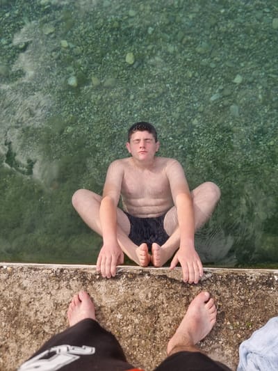 The image shows two people by a body of water with a concrete edge. One person, a young male, is partially submerged in the clear, rocky water, sitting cross-legged and looking upwards with his eyes closed. He is wearing black swim trunks. His arms are resting on his legs. The other person is on the concrete edge, with their feet visible. One foot is bare and pointing towards the water, while the other is covered by dark fabric. A light blue towel is draped on the concrete near the person's feet. The water appears to be shallow, with visible pebbles on the bottom. The lighting suggests it is daytime, and the overall impression is one of relaxation and enjoyment in a warm environment. The location is identified as Mosomishte, Bulgaria. There is no visible text in the image.