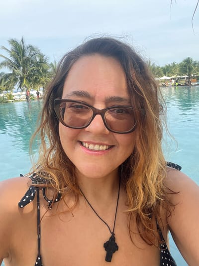 A woman takes a selfie by a swimming pool in Cam Lam, Vietnam. She is wearing a black and white polka dot bikini top and a dark necklace with a cross pendant. She has brown hair and is wearing glasses. The background shows palm trees, lounge chairs, and umbrellas by the pool.