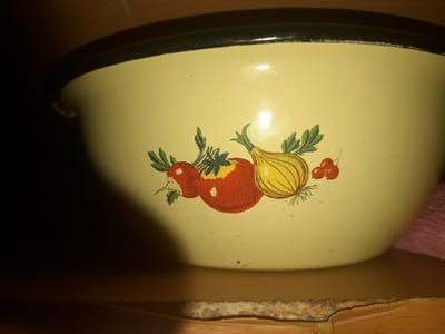 A close-up, low-angle interior shot shows a light yellow enamel bowl with a dark rim sitting on a wooden surface. The bowl features a colorful decal depicting a tomato, an onion, and cherries with green stems and leaves. The background is dark, with a slight glow on the upper right side, suggesting a light source outside the frame. The wooden surface appears worn and slightly damaged in the foreground. There are no people or animals visible in the image, and no specific geographical markers are present to indicate a location in Zilupe, Latvia. The lighting is dim, with a spotlight effect illuminating the bowl and a portion of the background.