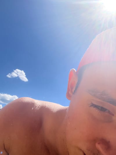 This is a close-up, low-angle outdoor photograph of a person in Delchevo, North Macedonia. The sky is a bright blue with a few scattered white clouds. The sun is shining intensely, creating a lens flare and highlighting the subject's skin, which appears to have water droplets on it, suggesting it is either wet or humid. The subject is wearing a red hat. The image focuses on the shoulder, ear, and a portion of the face, with the subject's eye closed. The overall impression is of a warm, sunny day.