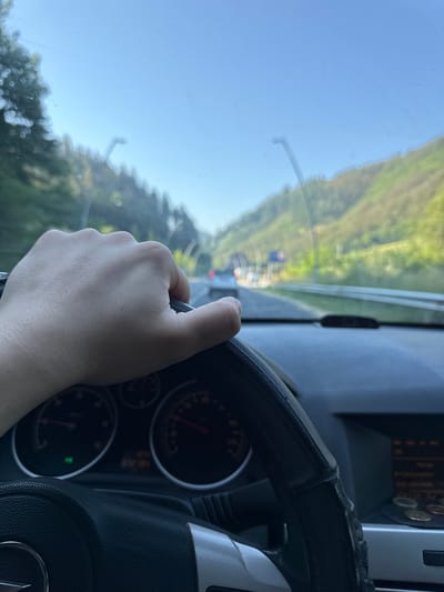 The image shows a person's hand on the steering wheel of a car. The car is driving on a road that appears to be in a mountainous area, with trees and green hills visible on either side. The sky is clear and blue. The dashboard of the car is visible, including the speedometer and other gauges. The image is taken from inside the car, looking forward. The location is identified as Madan, Bulgaria.