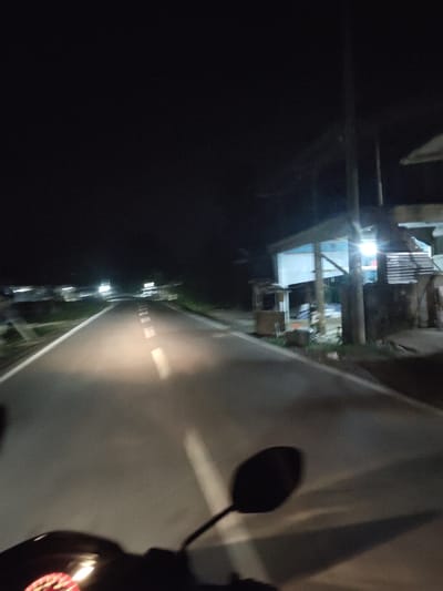 The image depicts a road at night, likely in or near Tanjongdrien, Indonesia. The perspective suggests the photo was taken from a moving vehicle, possibly a motorcycle given the presence of handlebars and a dashboard light in the foreground. The road is paved and has a white lane marker running down the center. Streetlights or lights from nearby structures illuminate the road, casting shadows and creating a somewhat blurry, dynamic scene. On the right side of the road, there appears to be a small building or stall with bright lighting, possibly a roadside shop or eatery. The background is dark, suggesting it's late at night. The overall impression is of a journey along a road at night in a semi-rural or suburban area.