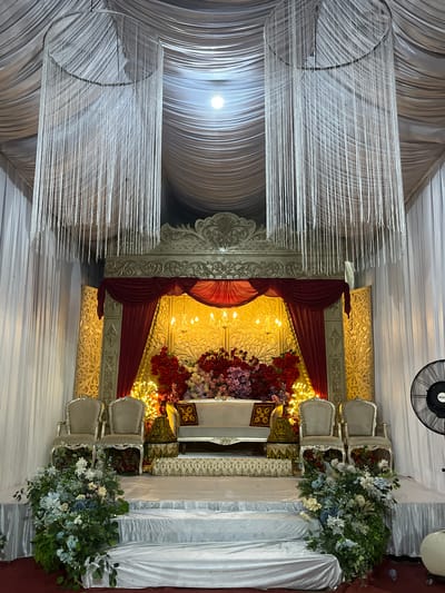 This media file captures a decorated stage set up for an event, possibly a wedding or a special ceremony, in Keudematangkuli, Indonesia. The scene is dominated by draped white fabric, creating a soft, elegant backdrop. Hanging from the ceiling are two large cylindrical chandeliers made of numerous white strands, adding a shimmering, ethereal quality to the space.
The focal point of the stage is an ornate, gilded backdrop with intricate carvings. In front of this backdrop, red velvet drapes are strategically placed, adding a touch of luxury and drama. The stage is adorned with vibrant floral arrangements, featuring a mix of red and pink flowers, along with softer blues and whites, creating a striking contrast against the rich red fabric.
Several chairs are arranged on the stage. Two chairs, upholstered in a light color and featuring ornate silver frames, are positioned prominently. In front of them, a white table is set, suggesting a place of honor. The lighting appears to be warm and ambient, with strings of small, yellow lights woven through the floral displays, enhancing the festive and celebratory atmosphere.
To the upper right of the image, a black industrial fan is visible, suggesting the need for ventilation in the enclosed space. The overall impression is one of a well-decorated and prepared venue for a significant occasion. There is no visible text within the image.