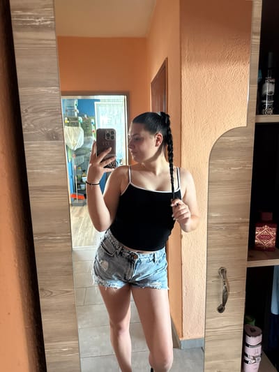 A young woman stands in front of a mirror, taking a selfie. She is wearing a black tank top and denim shorts. Her hair is pulled back in a braid. The mirror reflects a room with orange walls and a wooden cabinet. A shelf in the cabinet holds several bottles of perfume. The overall scene appears to be an indoor setting, likely a bedroom or dressing area. The location is specified as Rogosh, Bulgaria.