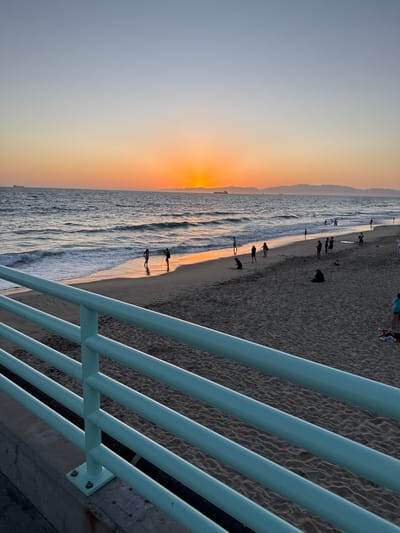 This image captures a sunset scene at Manhattan Beach, California, USA. The foreground is dominated by a pale blue railing that runs diagonally across the frame. Beyond the railing, a sandy beach stretches out, dotted with several people. Some individuals are walking along the shoreline, while others are sitting or standing on the sand. The ocean is visible in the midground, with gentle waves lapping at the shore and reflecting the warm hues of the sunset. Several ships are visible on the horizon, and a mountain range can be seen in the far distance. The sky transitions from a pale blue at the top of the frame to a vibrant orange and yellow near the horizon where the sun is setting. The overall atmosphere is serene and tranquil.