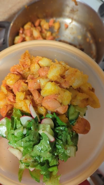 The image presents a close-up, high-angle view of a meal contained within a light orange plastic bowl. The bowl is divided visually into two primary food components. The upper section consists of cooked, diced potatoes intermixed with irregular slices of frankfurter-style sausages, all coated in a vibrant orange-yellow spice mixture. The lower section of the bowl holds a green salad, primarily composed of chopped lettuce leaves, multiple thin slices of radish displaying white interiors and pink edges, and visible green cucumber slices. In the blurred background, a metallic pot, likely stainless steel, contains additional quantities of the potato and sausage mixture, indicating a food preparation or serving environment. No individuals, actions, or specific interactions are depicted. The precise location within Modiin-Maccabim-Reut, Israel, cannot be determined from the visual content of the image, which solely focuses on the food and immediate surroundings consistent with a domestic kitchen or dining setting.
