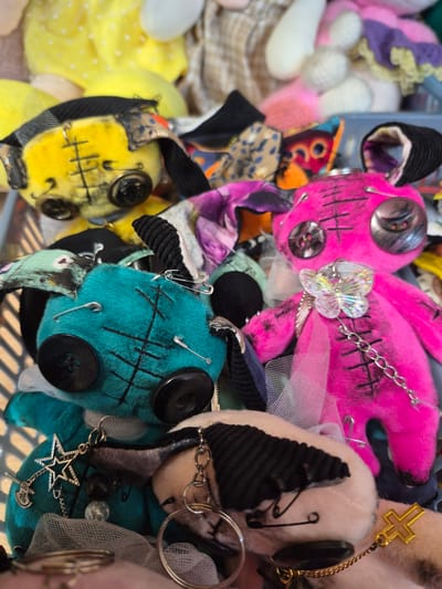 The image displays a collection of handmade stuffed toys, possibly in a craft market or workshop setting in the city of Krasnyy Klyuch, Russia. The toys are eclectic in design, with a focus on a "creepy-cute" aesthetic.
One prominent toy is bright pink, with large black buttons for eyes and stitched details. It is adorned with a delicate iridescent butterfly charm and a silver chain. Another toy, positioned to the left and slightly behind the pink one, is teal-colored, also featuring button eyes and stitched markings. It has safety pins attached to its body. A yellow toy, partially visible in the upper left, shares similar button eyes and stitched features, with a black and patterned fabric headpiece. Other toys are visible in the background, some appearing to be made of pastel-colored yarn or fuzzy material.
The toys are arranged in what appears to be a basket or container, with various fabric scraps and embellishments scattered around them. The lighting suggests an indoor setting, possibly with natural light filtering in. The overall impression is one of artisanal crafting, with a focus on unique, characterful creations.