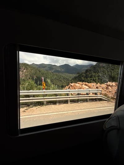This media file captures a scenic view from a window, likely of a vehicle, looking out onto a mountainous landscape. The frame of the window is dark, contrasting with the brightly lit exterior. The scene is oriented vertically in the image, suggesting it was taken with the camera rotated.
The main subjects are the natural environment and man-made structures along a road. A winding road is visible, bordered by a metal guardrail. Beyond the road, a steep, rocky slope covered in dense green trees dominates the left side of the frame. In the distance, rolling mountains fade into a cloudy sky. A traffic sign, yellow with a black arrow indicating a sharp turn, is prominently placed near the roadside vegetation.
The time of day appears to be late afternoon, with long shadows cast by the sun hitting the mountains and road. The weather seems partly cloudy, with sunlight breaking through. The location, Nam Khanh Vinh, Vietnam, is hinted at by the lush, tropical vegetation and mountainous terrain. There are no people visible in the frame, and the overall mood is one of natural beauty and peaceful travel. The only visible text is on the traffic sign, which is a directional arrow.