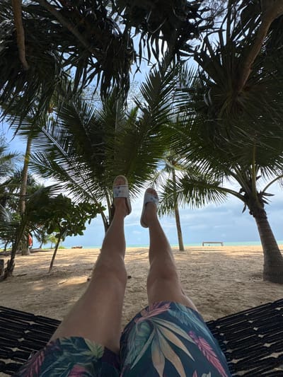 A first-person perspective shot shows a person relaxing in a hammock on a sandy beach, likely in Ban Khuk Khak, Thailand. The person's legs are extended, wearing floral-patterned shorts and light-colored flip-flops. Palm trees with lush green fronds frame the scene from above, casting shadows on the sand. In the background, the turquoise ocean meets a cloudy sky. The beach appears uncrowded, with a wooden structure or bench visible in the distance near the water. The overall atmosphere is one of tropical tranquility.