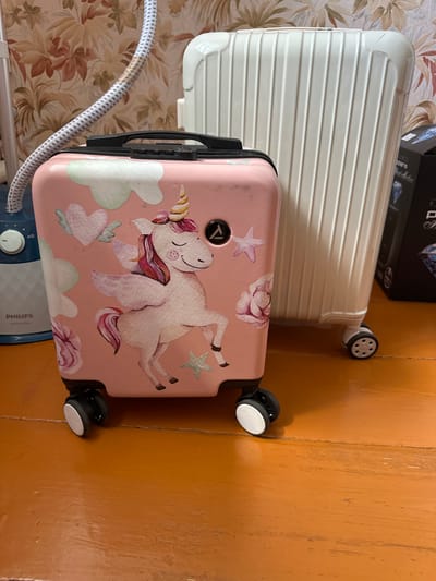 The image shows a pink suitcase with a unicorn decal and a white ribbed suitcase standing on an orange wooden floor. A garment steamer is partially visible to the left of the pink suitcase, and a black box with a diamond graphic is to the right of the white suitcase. The background features floral wallpaper. The scene is indoors and appears to be in a residential setting. There are no people visible in the image. The location is described as Urazovo, Russia, though there is no visual information within the image to confirm this specific location.