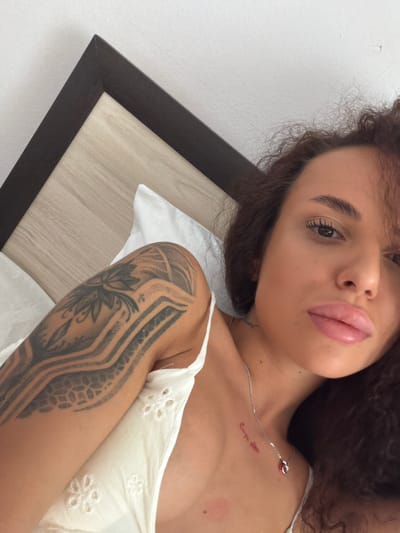 A young woman with curly dark brown hair and a floral tattoo on her left arm lies in bed. She is wearing a white top. Her face is partially visible, with her eyes closed and her lips slightly parted. The background shows a wooden headboard. There is no indication of the location in the city of Petrich, Bulgaria.