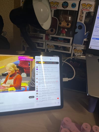 This image captures an indoor desk setup in San José Pinula, Guatemala. A black flexible-neck desk lamp with a white bulb is positioned above a tablet, which is actively displaying a live video stream alongside a scrolling chat window. The video feed shows a light-skinned woman with blonde hair, wearing an orange overall-style apron over a dark long-sleeved shirt, preparing what appears to be raw meat on a yellow cloth. The chat window, labeled "Chat en vivo" (Live chat), displays numerous comments in Spanish, including critical remarks such as "ELEAZAR LÁRGATE" (Eleazar get out) and "FUERA PATRON DA HUEVAS" (Out with the annoying boss/patron). A pink banner at the top of the tablet screen advertises "Disney+ | ¡Suscríbete!". Behind the tablet and lamp, multiple Funko Pop figures in their original packaging are stacked, including "MAC" (#941) and "AGATHA HARKNESS." To the right, a silver laptop is open, displaying what appears to be an inventory or data management interface with English text. A white cable connects to the right side of the tablet, extending towards the laptop area. In the bottom right corner, a partial view of a pink and white mechanical keyboard is visible. The desk surface is light-colored, and the background wall is a plain light hue.
