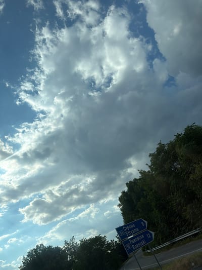 The image depicts a rural roadside scene under a dramatic sky. Two blue directional signs are prominently displayed, indicating "Струмя Stryama 8" and "Раковски Rakovski 12", with the number 56 on both. The signs are positioned at a junction of two roads, one of which curves out of frame to the right. Lush green trees line the roadside, forming a dense canopy that partially obscures the sky. The sky itself is filled with a mix of light and dark clouds, with patches of blue sky visible, suggesting a dynamic weather pattern. The overall impression is one of a scenic, perhaps somewhat remote, location. Based on the directional signs, the location is Skutare, Bulgaria.
