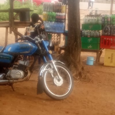 The image captures an outdoor scene in Gumel, Nigeria, likely during the daytime given the lighting. The foreground is dominated by a blue motorcycle, parked on a dirt ground. The motorcycle appears to be in use, with visible chrome details and a well-worn engine. A person, partially visible and wearing a patterned shirt, is seated on the motorcycle, with their hand raised to their head, possibly indicating a moment of rest or contemplation.
Behind the motorcycle, a wooden structure serves as a makeshift stall or vendor stand. On this stand and the ground next to it, there are stacks of beverages, predominantly bottled drinks. These include cases of what appear to be soft drinks, with visible labels and colored bottles (green, blue, and yellow). One red crate clearly displays the "Coca-Cola" logo. A yellow crate on the right side also has visible text, including "GUINNESS." A blue cooler is placed on the ground near the stall.
In the background, there is lush green vegetation, suggesting an area with some natural elements. The overall setting appears to be a roadside stall or a local market area. The presence of the motorcycle and the stacked beverages indicates a commercial activity, likely the sale of drinks. The image has a slightly grainy quality, characteristic of older or lower-resolution digital photography. The ground is sandy or dusty, consistent with arid or semi-arid environments.