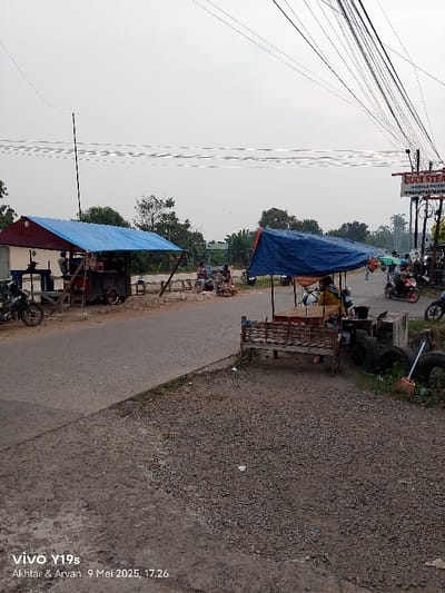 The image shows a street scene in Kecamatan Kroya, Indonesia. It's a sunny day with a partly cloudy sky. The image is taken from a perspective on the side of a paved road, looking towards a couple of street vendors. The vendors have makeshift stalls under blue tarps, and there are several people sitting at the stalls. There are also a few motorbikes parked in the street, and a few people walking around. The street is lined with gravel on the sides. There is a sign on the right side of the image that says "GUCI STRA", and there is a sign on the left side of the image that is not visible in the image. The image is taken from a low angle, giving the viewer a perspective of the street scene. The image was captured on a smartphone. The image is dated May 9, 2023 at 5:26 pm.