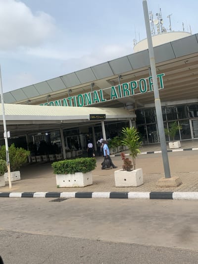 The image captures the entrance area of an airport building, identified by the prominent green lettering spelling "INTERNATIONAL AIRPORT" on its facade. The building features a modern design with large glass windows and a wide canopy over the entrance.
Below the main sign, a smaller white sign with arrows indicates "Domestic Departure" and "International Departure." Several individuals are visible near the entrance, suggesting typical airport activity. One person in a light blue shirt and dark trousers is standing next to a concrete planter, carrying a black backpack. Other people in white shirts can be seen further inside the entrance.
The foreground shows a paved area, likely a pedestrian walkway or drop-off zone, bordered by a black and white striped curb. Concrete planters filled with green shrubs and smaller plants are positioned along the building's edge. A plain grey pole, possibly a signpost or light post, stands vertically in the mid-ground.
Above the airport building, against a partly cloudy sky, a collection of antennas and satellite dishes is visible on a rooftop or tower, indicative of communication infrastructure common at airports. The scene is brightly lit, suggesting daytime conditions. The setting is an international airport in Gwaska, Nigeria.