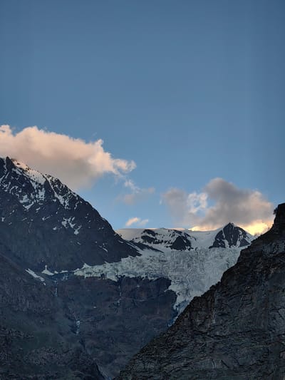 A vertical, medium shot captures a majestic mountain landscape under a partly cloudy sky. The sky transitions from a deep blue at the top to a lighter shade near the horizon. Puffy, white clouds, tinged with peach and gold from the setting sun, drift across the sky, partially obscuring the rugged mountain peaks.
The mountains themselves are a dramatic contrast of dark, rocky slopes and dazzling white snow. The snow caps the higher elevations, creating sharp, jagged silhouettes against the sky. A prominent glacier, a mass of textured ice, cascades down the face of one of the mountains, its surface marked by deep crevasses and icy ridges. Water appears to be flowing down the rocky slopes in thin streams, hinting at the melting ice.
In the foreground, a dark, craggy mountain face dominates the right side of the frame, its rough texture adding depth to the scene. A small, lone tree clings precariously to the upper edge of this rock face. The overall impression is one of raw, natural beauty and the immense scale of the alpine environment. The warm glow on the clouds suggests that it is either sunrise or sunset, casting a serene and dramatic light on the scene. The location context provided is Kead, India, which is a mountainous region. No people, objects, or text are visible in the image.