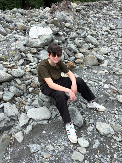 An outdoor scene in Lagodekhi, Georgia, depicts a young male seated on a large, light-grey rock within a rocky, riverbed-like environment. The individual wears dark-tinted sunglasses, a dark olive green short-sleeved t-shirt with a subtle front graphic, black trousers, and white sneakers with red and green stripes, complemented by white socks with black stripes. A light-colored, possibly braided, bracelet is visible on his left wrist. He is positioned facing the viewer with a slight smile.
The ground surface is predominantly composed of numerous grey rocks of varying sizes and shapes, interspersed with darker gravel and sand. A light-colored, folded item of clothing, possibly a t-shirt or towel with a partial graphic, is resting on a prominent rock behind and to the left of the individual. Scattered dry branches and twigs are visible among the rocks, particularly in the mid-ground and background. Dense green foliage, characteristic of trees or bushes, forms the horizon line in the far background. A minimal amount of water, suggesting a narrow stream or pooling, is discernible in the far right middle ground. The lighting is diffused, indicating an overcast sky or shaded conditions.