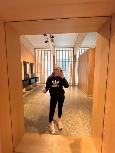 A young female stands centered in the reflection of a large, light-wood framed mirror, taking a selfie with a smartphone. The scene is located inside the Visitor Centre at the Gotthard Base Tunnel in Erstfeld, Switzerland. The female wears a black hooded sweatshirt with a white Adidas trefoil logo, black leggings, white and orange athletic shoes, and pink ankle socks. Her dark hair is pulled back, and her mouth is slightly open.
The reflected background reveals a modern, spacious interior with light-toned wooden wall panels, a polished concrete floor, and distinctive vertical wooden slat structures serving as large window coverings, allowing diffused light to enter. On the left side of the reflection, two dark, rectangular wall-mounted sink units with individual rectangular mirrors above them are visible, indicating a public washroom area. The ceiling features light panels, exposed dark track lighting fixtures, and light-colored beams. On the right, a section of a light wood partition wall is reflected. The overall environment exhibits a clean, contemporary architectural design with natural materials.