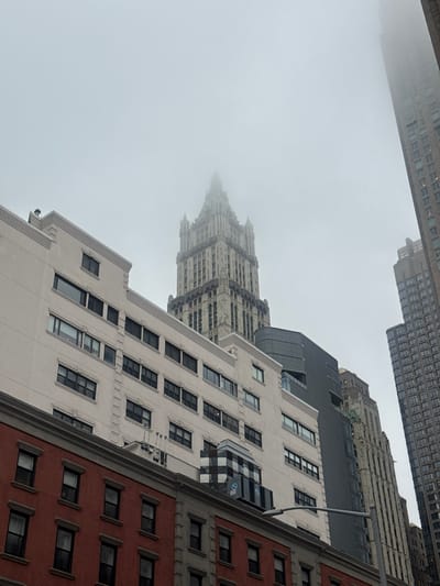 The image captures a view of urban architecture in Hoboken, United States of America. The foreground is dominated by the upper floors of two buildings. The building on the far left is a red brick structure with multiple evenly spaced windows. Above it, a white building with a textured facade displays rows of dark-framed windows. Further back and to the right, a series of taller, modern buildings rise into a foggy sky. One prominent building in the middle distance features a distinctive Gothic-inspired spire, partially obscured by fog. Another tall, rectangular skyscraper with a grid of windows is visible on the far right, also fading into the mist. The overall atmosphere is overcast and hazy.