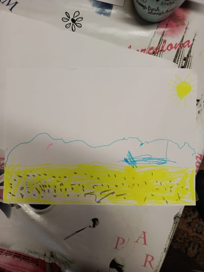 The image shows a child's drawing on a piece of white paper. The drawing depicts a landscape with a yellow field, a blue outline of hills or mountains, and a yellow sun in the upper right corner. There are also some dark speckles scattered across the yellow field, possibly representing flowers or stones. A small pink zig-zag line is present on the blue hills. The paper is placed on a table with a patterned surface. There is no information in the image that indicates the location in the city of Meget, Russia.