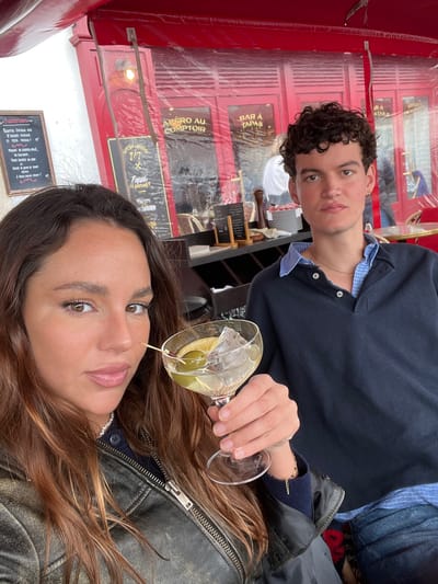A selfie shows two individuals at an outdoor cafe in Bayonne, France. The person taking the photo is a woman with long brown hair, wearing a black leather jacket and a pearl necklace. She is holding a martini glass with a toothpick skewering olives. Behind her sits a man with dark curly hair, wearing a navy blue polo shirt and denim jeans. The background reveals a red structure with transparent plastic panels, bearing the words "BAR A TAPAS," "APERO AU COMPTOIR," and "Tapal a partage." A chalkboard menu displays "SUGGESTIONS" in French.