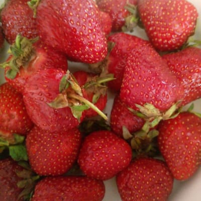 The image displays a close-up, top-down view of a pile of fresh, ripe strawberries. The strawberries are a vibrant red color, with visible seeds dotting their surfaces. Some of the strawberries have green leaves or stems attached. The lighting is bright, suggesting it is daytime, and the focus is sharp, highlighting the texture of the fruit. The setting appears to be a bowl or container, though it is not fully visible. The image does not contain any people, text, or discernable location cues beyond the subject matter itself. The overall impression is one of freshness and abundance of fruit. The location context provided is Ashtarak, Armenia, but this information cannot be visually confirmed from the image alone.