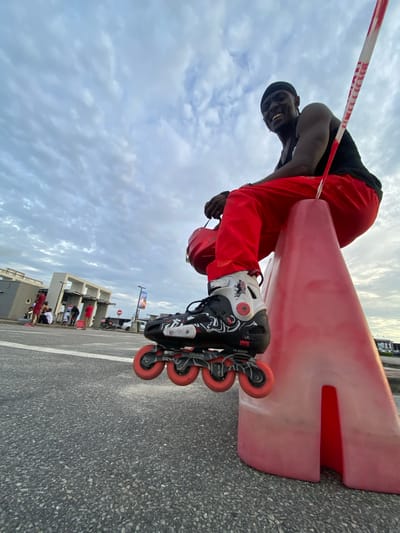 A low-angle, rotated view shows a man in red pants and roller skates standing on an asphalt surface next to a red and white traffic barrier, likely in an urban setting. The man is smiling, and a red and white caution tape crosses the upper left of the frame. In the background, a building and some people are visible under a cloudy sky. Based on visual cues, this could be Sangotedo, Nigeria.