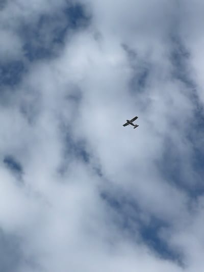 A small, single-engine airplane is captured in flight against a cloudy sky. The aircraft, silhouetted in dark tones, is positioned slightly off-center in the frame, appearing to bank gently. Its wings are extended, and the tail structure is clearly visible. The sky is filled with a mix of white, fluffy clouds and darker, more ominous patches, suggesting a dynamic weather system. The blue of the sky is visible in the gaps between the clouds, hinting at daylight, likely mid-morning or afternoon, given the illumination. No people are visible. The overall scene evokes a sense of freedom and exploration, with the vastness of the sky and the solitary presence of the airplane. No text is present in the image. The setting is likely aerial, with the airplane soaring over or within a cloudy atmosphere, possibly over the Gauja region of Latvia.