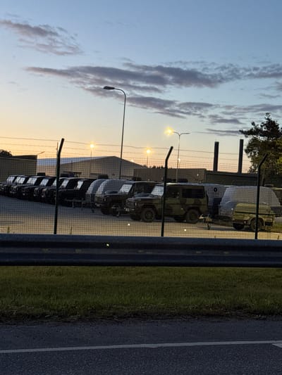 The image captures a fleet of dark-colored military vehicles parked in rows behind a wire fence, possibly in a secure compound. The sky above displays a gradient of light blue with scattered clouds, suggesting it is either dawn or dusk. Several streetlights are visible, some illuminated, casting a warm glow. In the background, buildings and trees form the horizon. A chain-link fence with barbed wire runs horizontally across the middle ground. The foreground shows a dark road and a strip of green grass. One of the vehicles, a camouflage-patterned jeep, is hitched to a trailer. The scene suggests a military or government facility in Paldiski, Estonia.