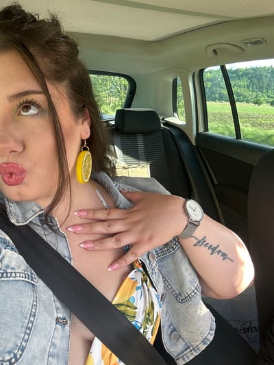 A woman with brown hair, hazel eyes, and light skin is sitting in a car, wearing a light blue denim jacket over a floral print top, and a black seatbelt. She has a lemon slice earring in her left ear, pink lipstick, and French-tip manicured nails. Her left arm is resting on her body and is visible, showing a watch on her wrist and a "freedom" tattoo on her forearm. The car is likely moving, as blurred greenery is visible through the open sunroof and side window. The location is inferred to be in or near Dragushinovo, Bulgaria, based on the prompt's request.