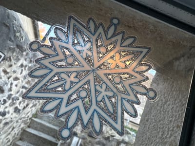 A decorative snowflake decal is affixed to the interior surface of a clear glass window pane. The snowflake design is multi-pointed, featuring white segments outlined in blue, with interior areas filled with textured silver glitter that reflects ambient light, displaying warm gold and orange tones. The decal is oriented slightly diagonally within the frame.
Through the window, a view of an outdoor urban environment in Le Puy-en-Velay, France, is visible. To the left and bottom-left, a robust stone wall constructed from dark, irregularly shaped stones is prominent, leading down to a visible section of stone steps. To the right and top-right, another rough-textured, lighter-colored stone facade is present. The dark frame of the window unit is visible along the top and right edges of the image. The scene is illuminated by daylight, with reflections evident on the glass. No people or active interactions are depicted.