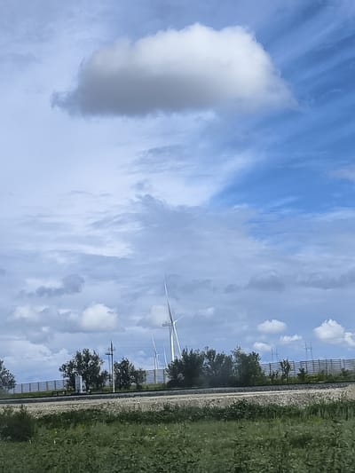 The image depicts a rural landscape with a cloudy sky. In the middle ground, several wind turbines are visible, some with their blades extended. A train track runs horizontally across the scene, with a fence and trees behind it. The foreground consists of dense green vegetation. The overall impression is one of an open, possibly agricultural area with a focus on renewable energy infrastructure. There are no people or animals visible in the image. The location is specified as Ақжар, Kazakhstan.